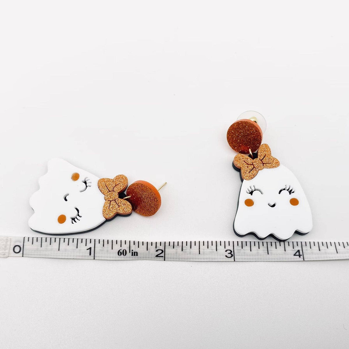 Pair of ghost-shaped earrings with bow details on a ruler for scale.