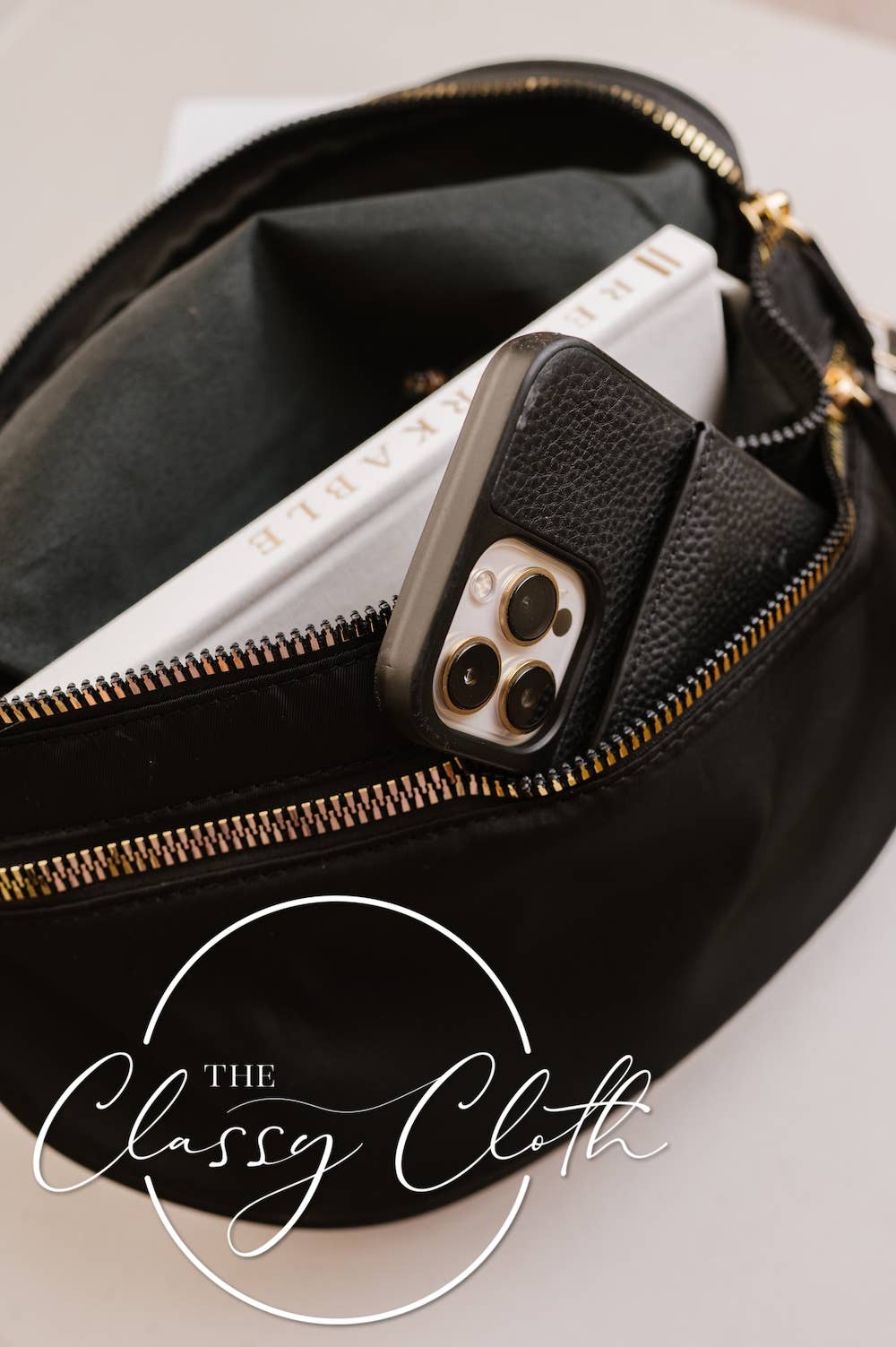 Black clutch bag with a phone and book inside, branded 'The Classy Cloth'.