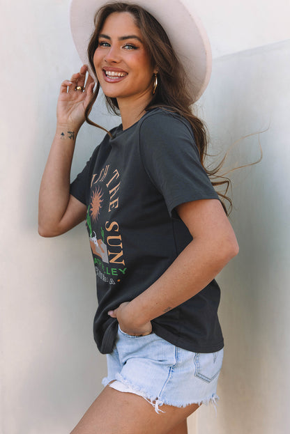 Woman wearing a black t-shirt with graphic design and denim shorts, standing against a light background.
