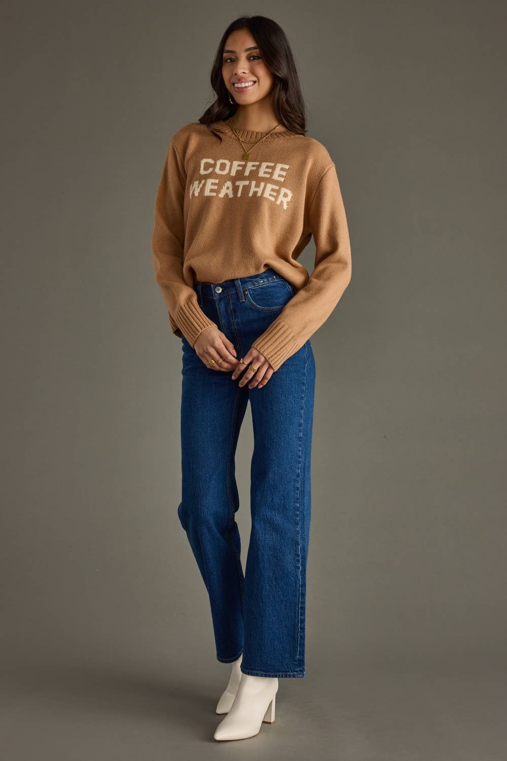 A woman modeling a tan crewneck sweater with 'COFFEE WEATHER' printed in white across the chest, paired with blue jeans and white ankle boots, against a solid gray studio background.