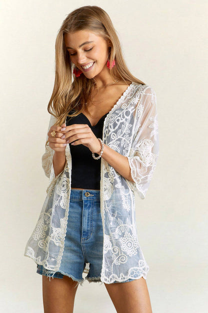 Woman wearing a lace kimono over a black top and denim shorts on a plain background