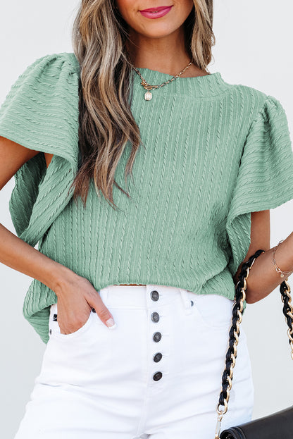 Woman wearing a green textured top and white pants with black buttons, holding a black handbag.
