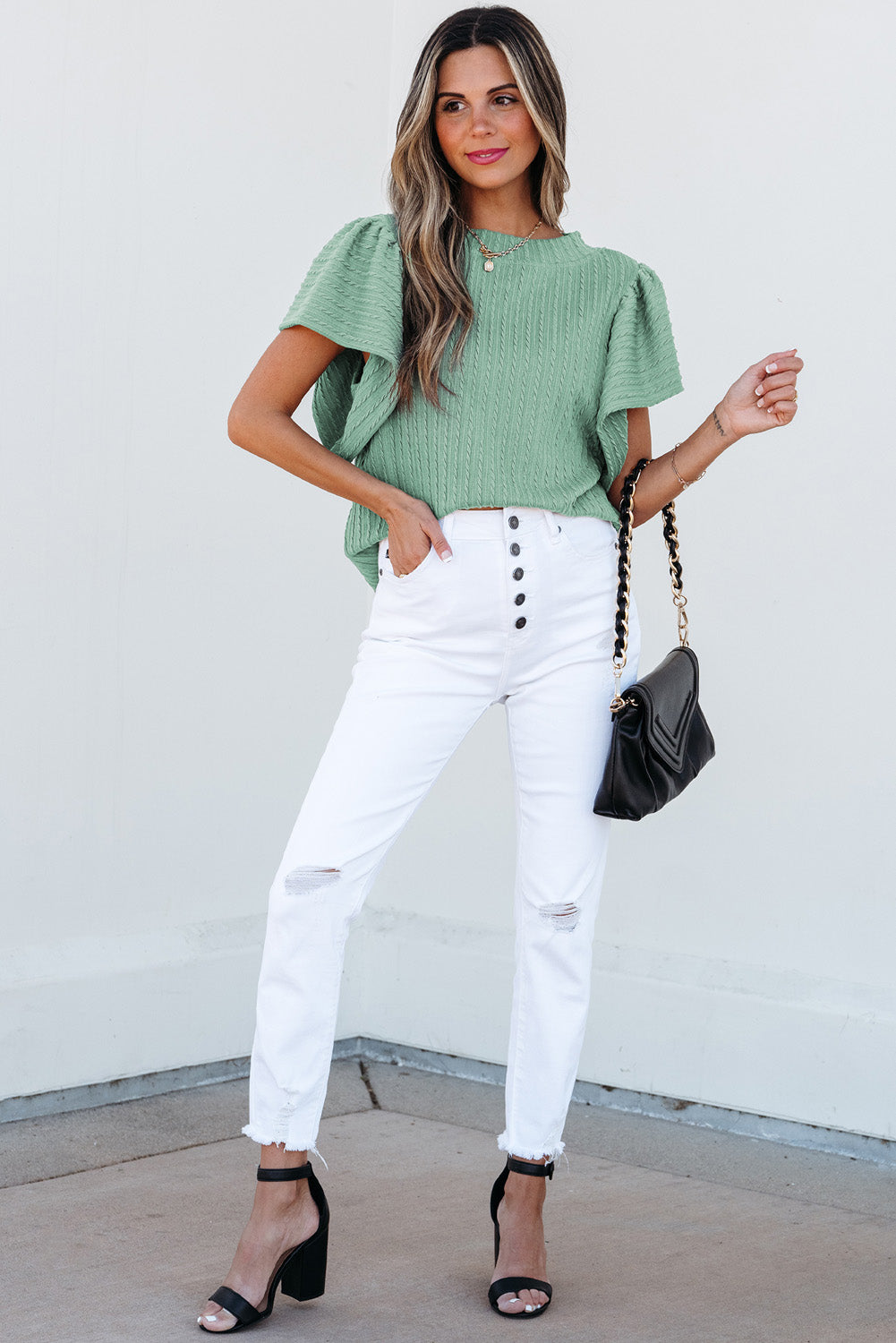 Woman wearing a green top and white pants with a black handbag against a white background