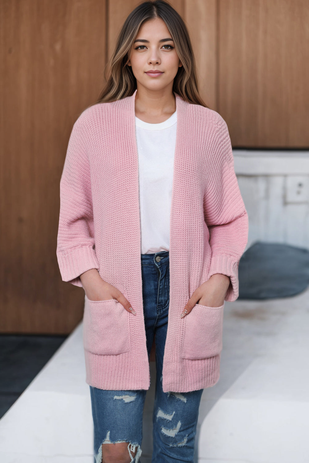 Woman wearing a pink cardigan over a white shirt and blue jeans, standing against a wooden wall.