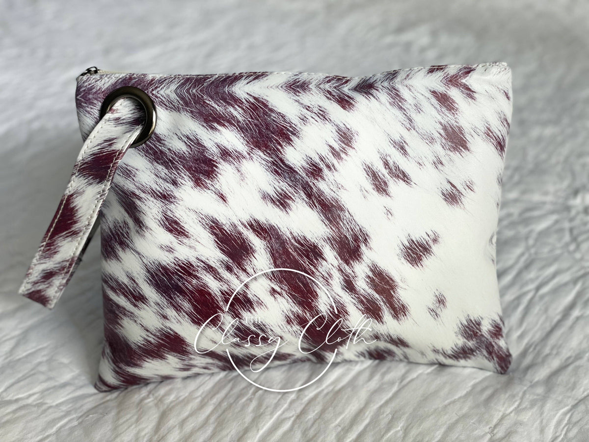 Clutch bag with maroon and white cow print pattern on a gray surface, featuring the brand ' classy cloth'.