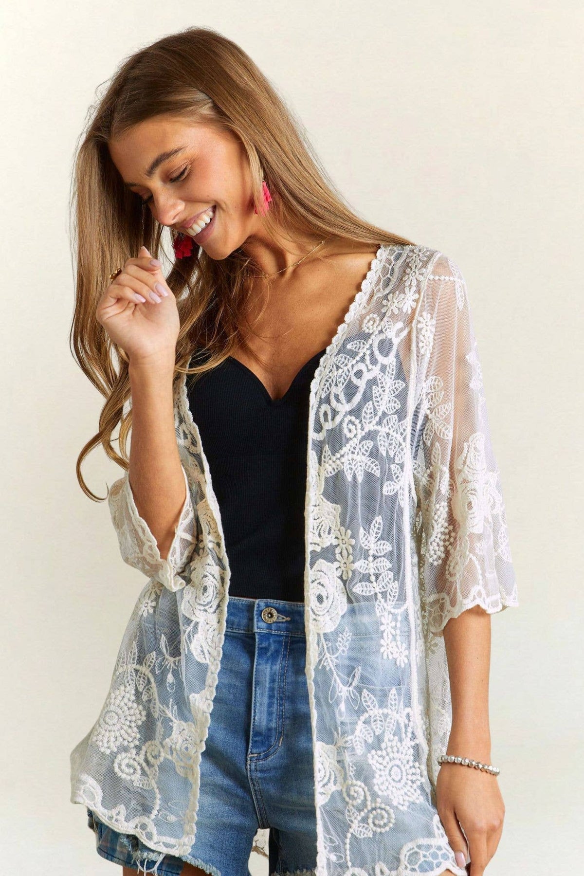 Woman wearing a lace kimono over a black top and denim shorts on a plain background