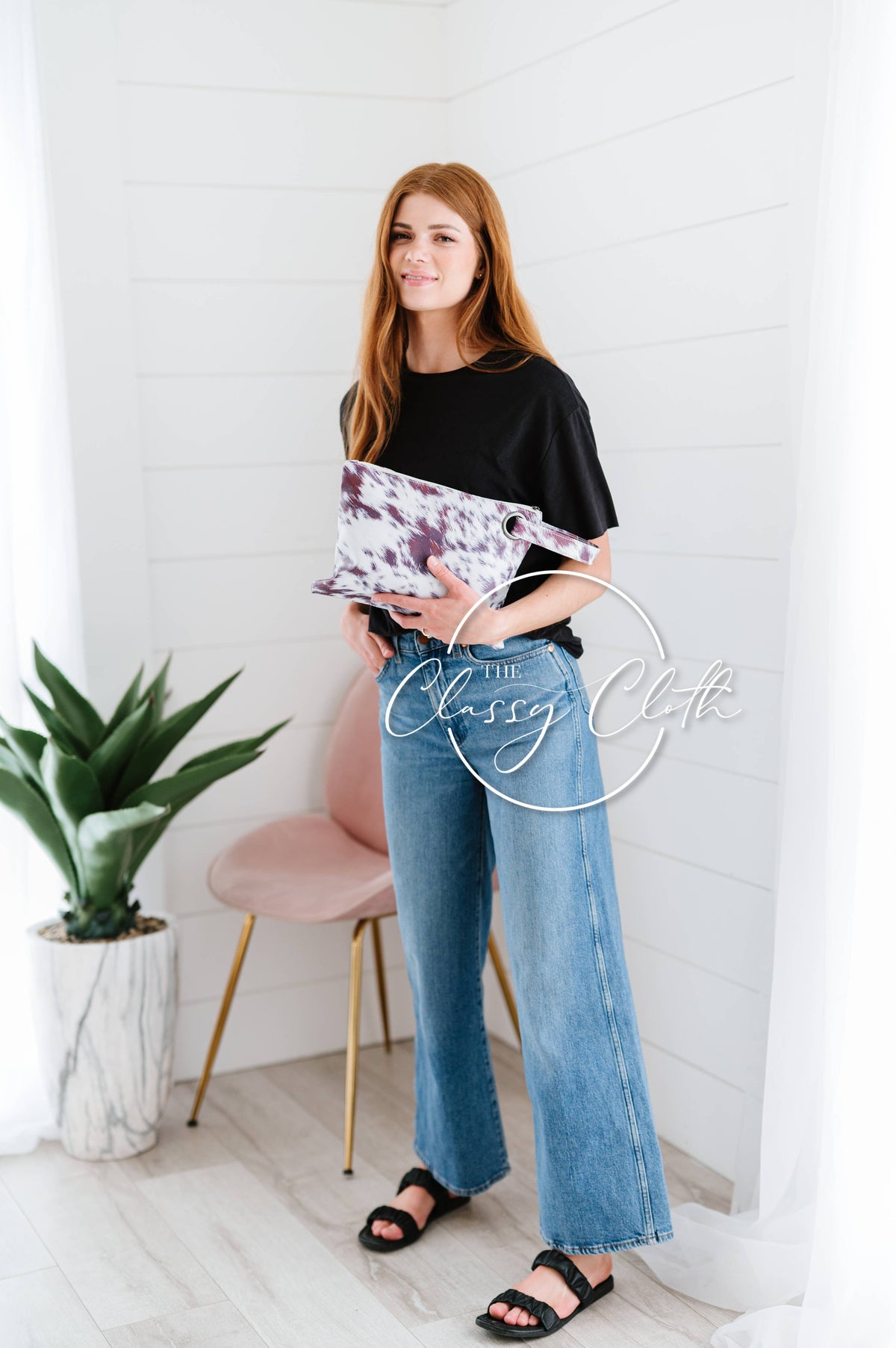Woman holding a clutch in a minimalistic room with a plant and chair.