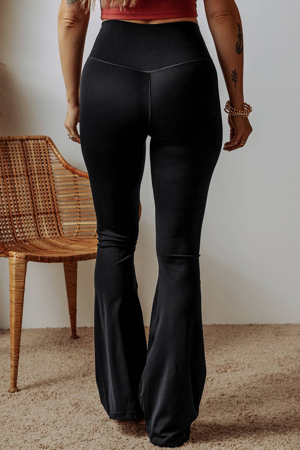 Person wearing black flared pants standing in a room with a wicker chair and beige carpet.