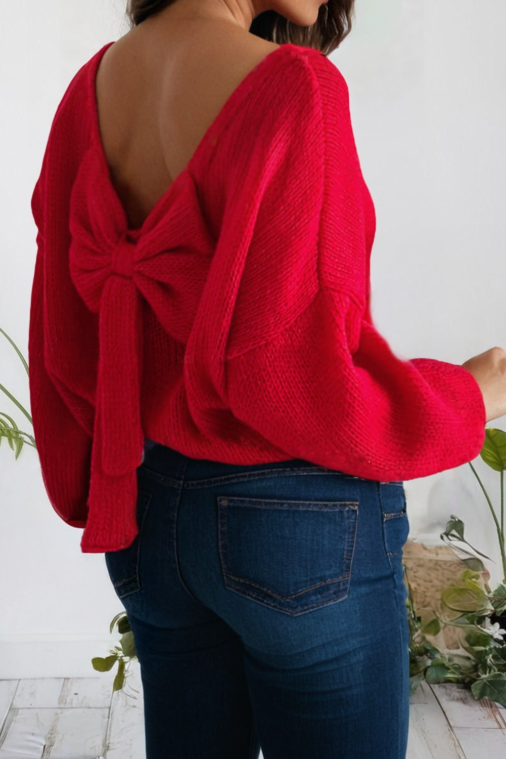 Person wearing a red sweater with an open back and blue jeans, standing against a white wall with plants.